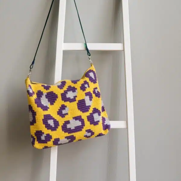 Keltainen ja violetti virkattu Leolaukku roikkuu valkoisilla tikkailla.