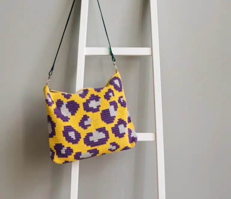 Keltainen ja violetti virkattu Leolaukku roikkuu valkoisilla tikkailla.