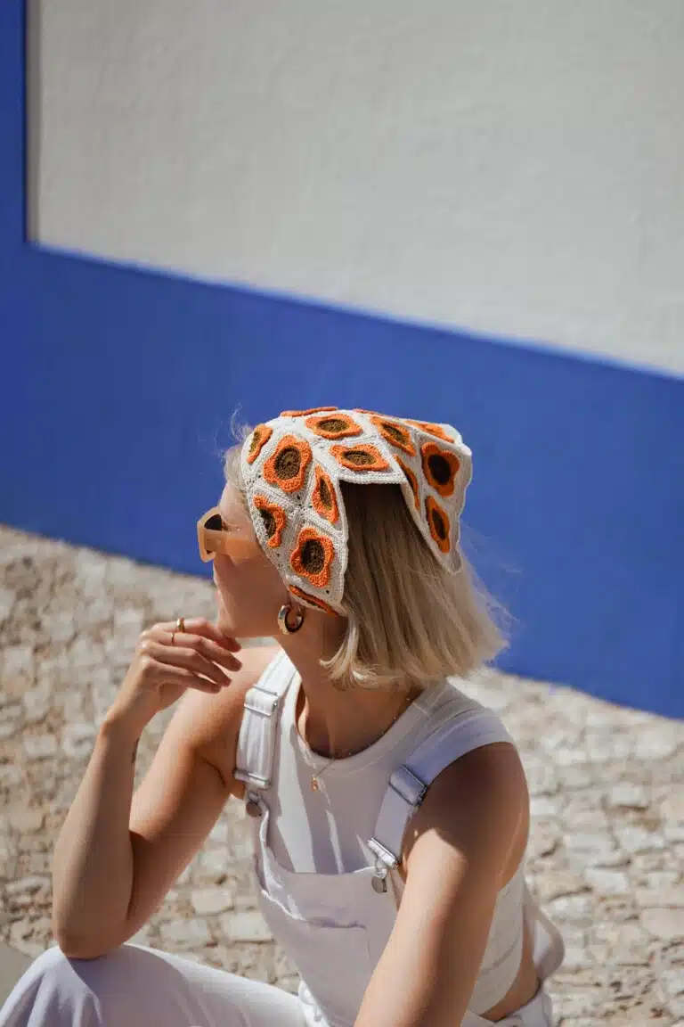 Nainen istuu aurinkoisella kivipinnalla sinistä seinää vasten, oranssikuvioinen Pirkko-bandana päässään.