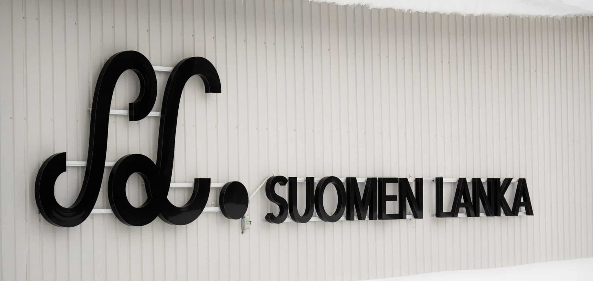 Suomen Langan musta logokyltti tehtaan valkoisessa seinässä.