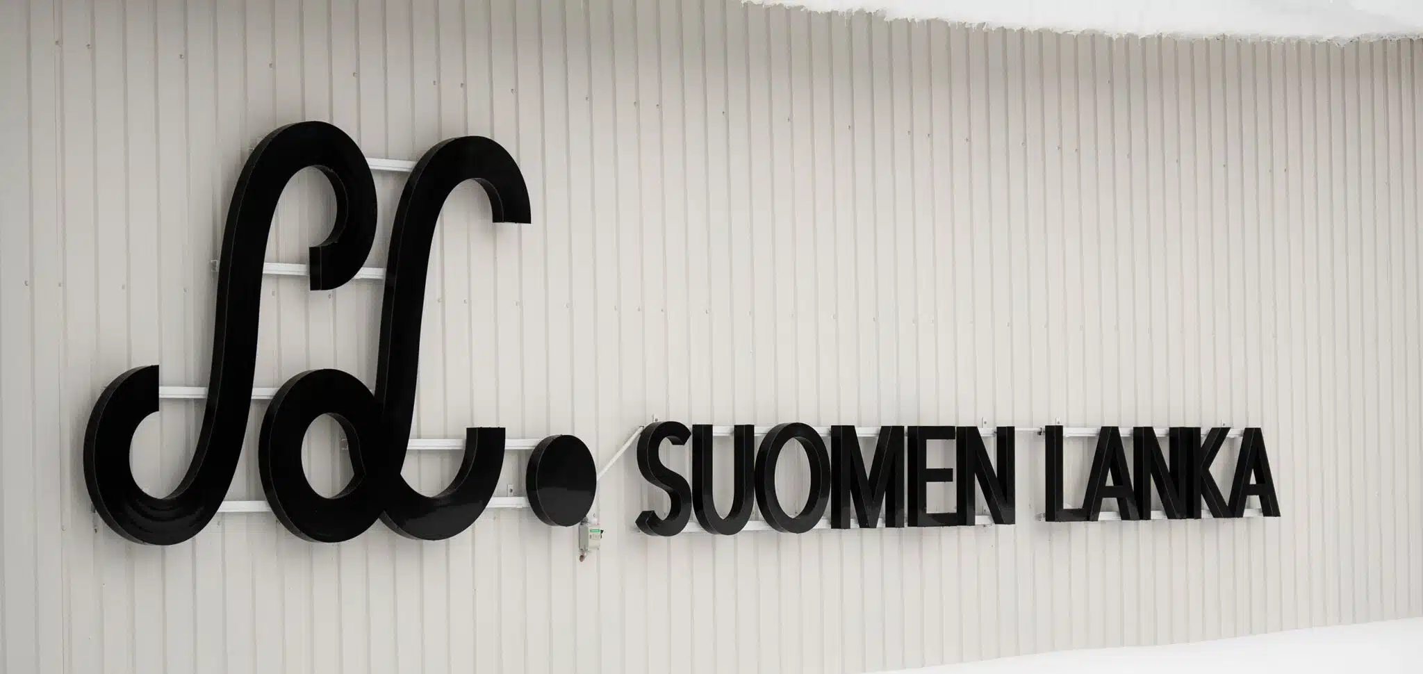 Suomen Langan musta logokyltti tehtaan valkoisessa seinässä.